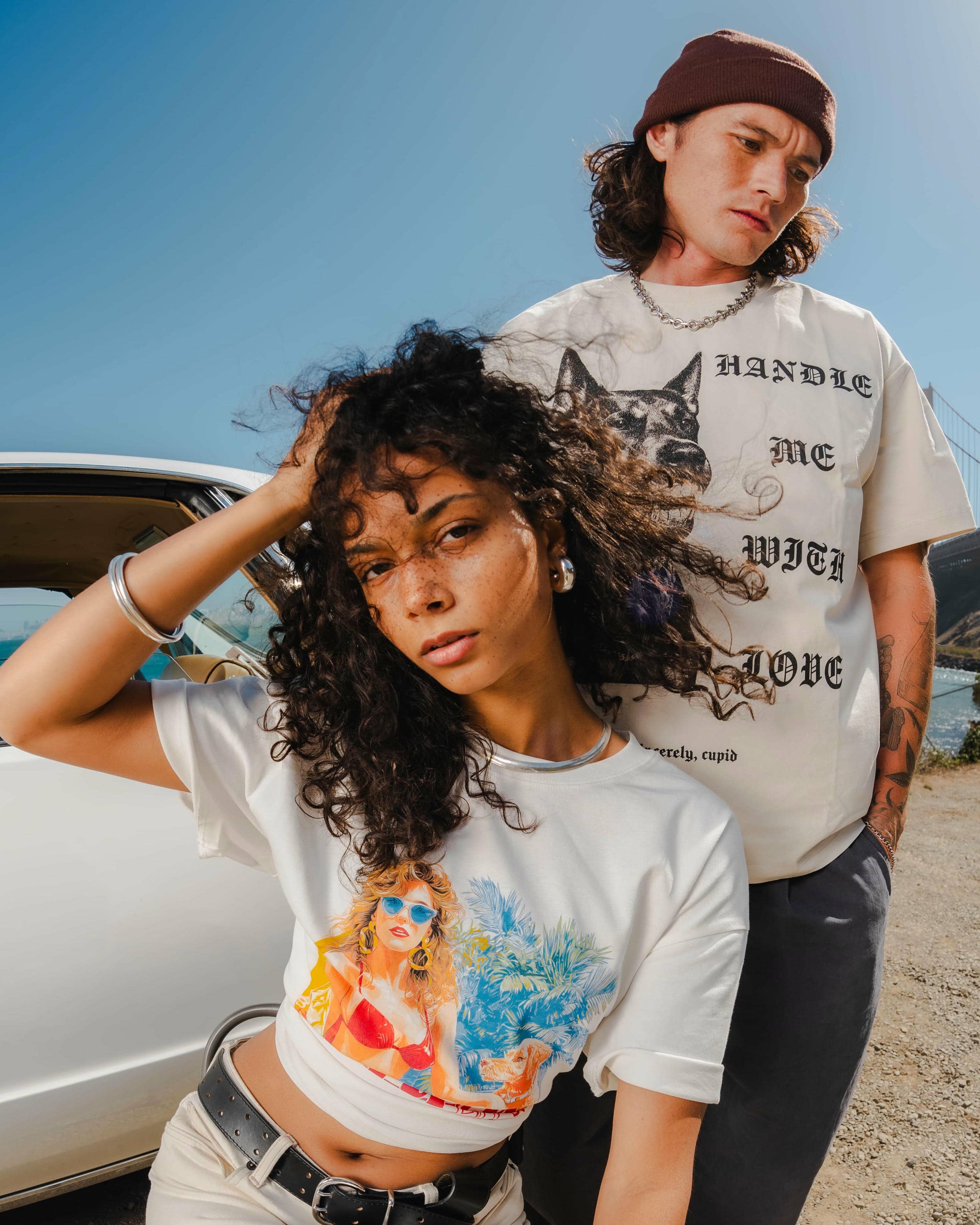 Two people wearing graphic t-shirts by a car with a scenic background