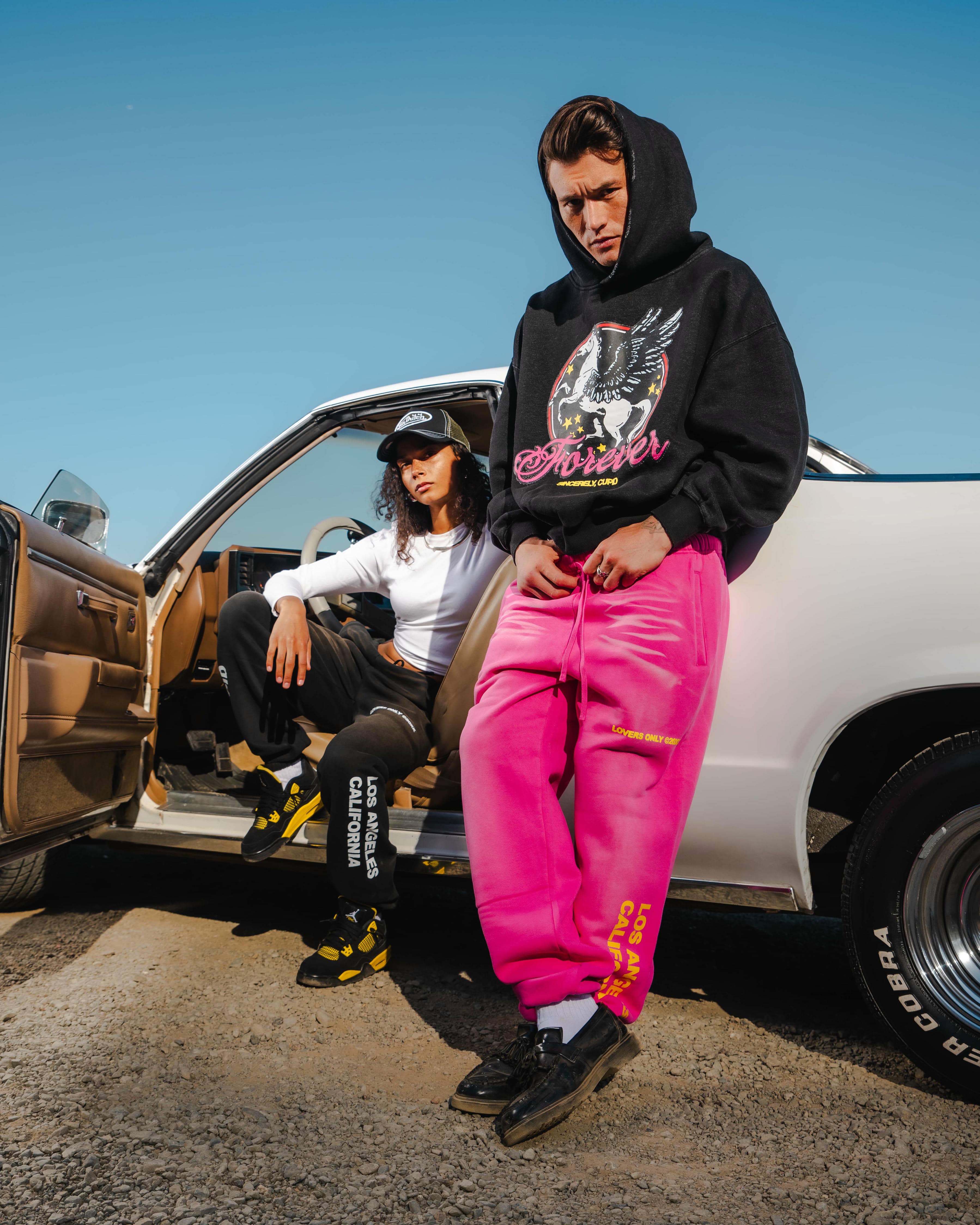 Two people in stylish outfits sitting by a classic car on a sunny day.